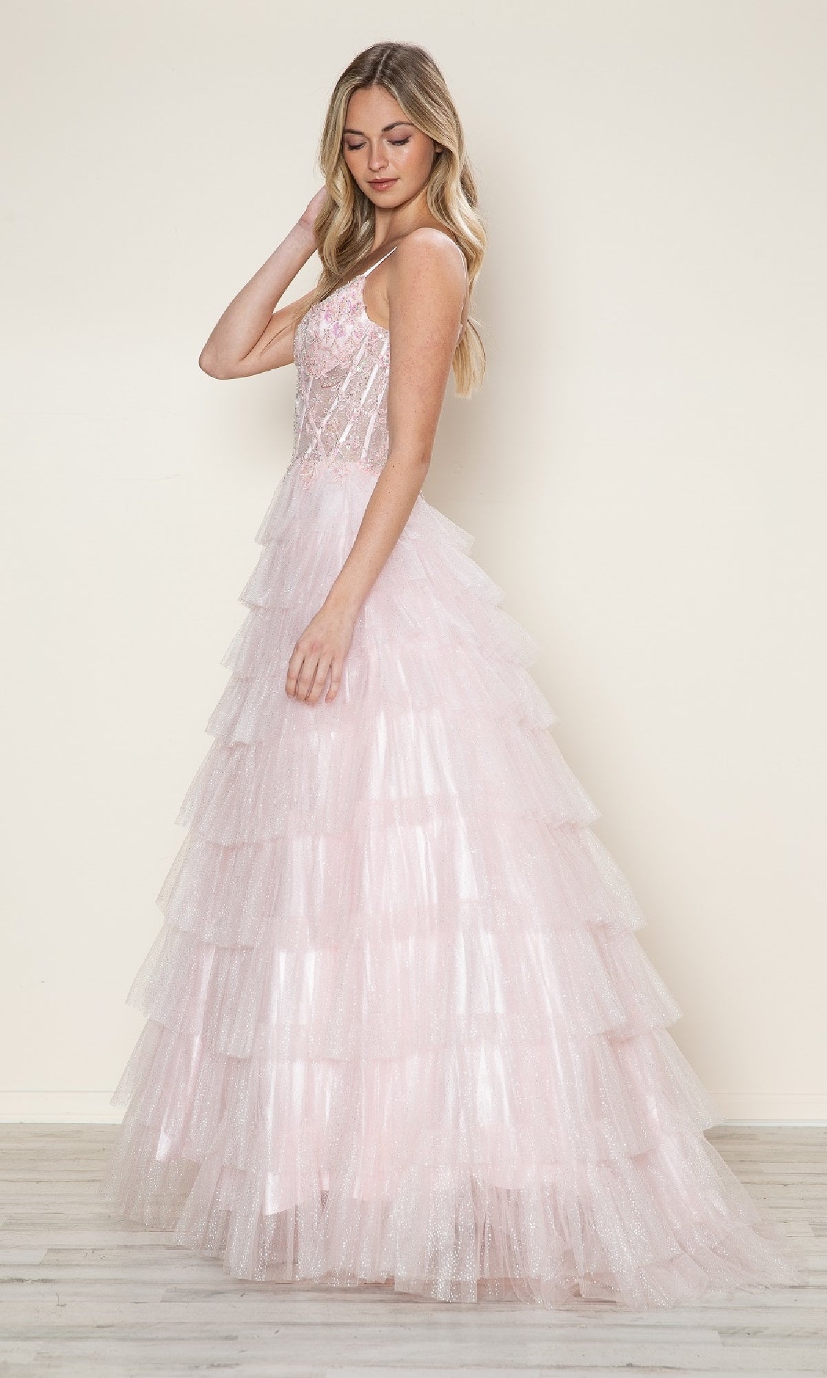 Woman wearing the Long Prom Dress: Poly USA 9608—a light pink, ruffled ball gown with a tiered skirt and spaghetti straps—stands sideways against a plain background, looking down.